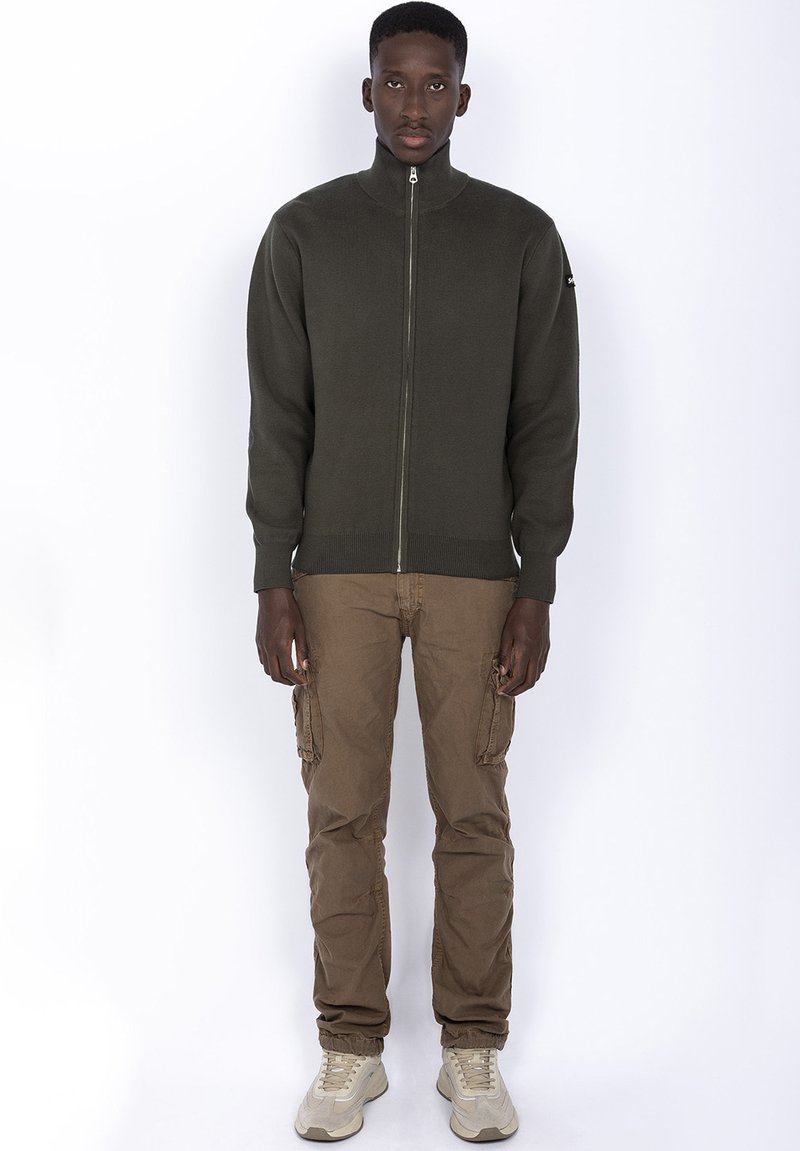 Mørkegrønt zip-up sweater med høj krave, båret over beige cargo bukser og beige sneakers. Stoffet ser glat og let struktureret ud.