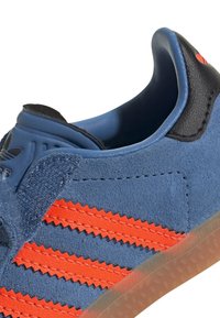 Blå mockasinsneaker med oranga zigzag-ränder, svarta detaljer och en genomskinlig gummisula. Rund tå och design med vadderad krage.