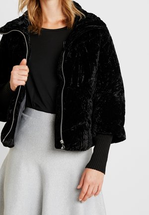 Kvinde iført en sort velour dunjakke over en sort top og en lysegrå tekstureret nederdel, stående foran en ensfarvet baggrund.