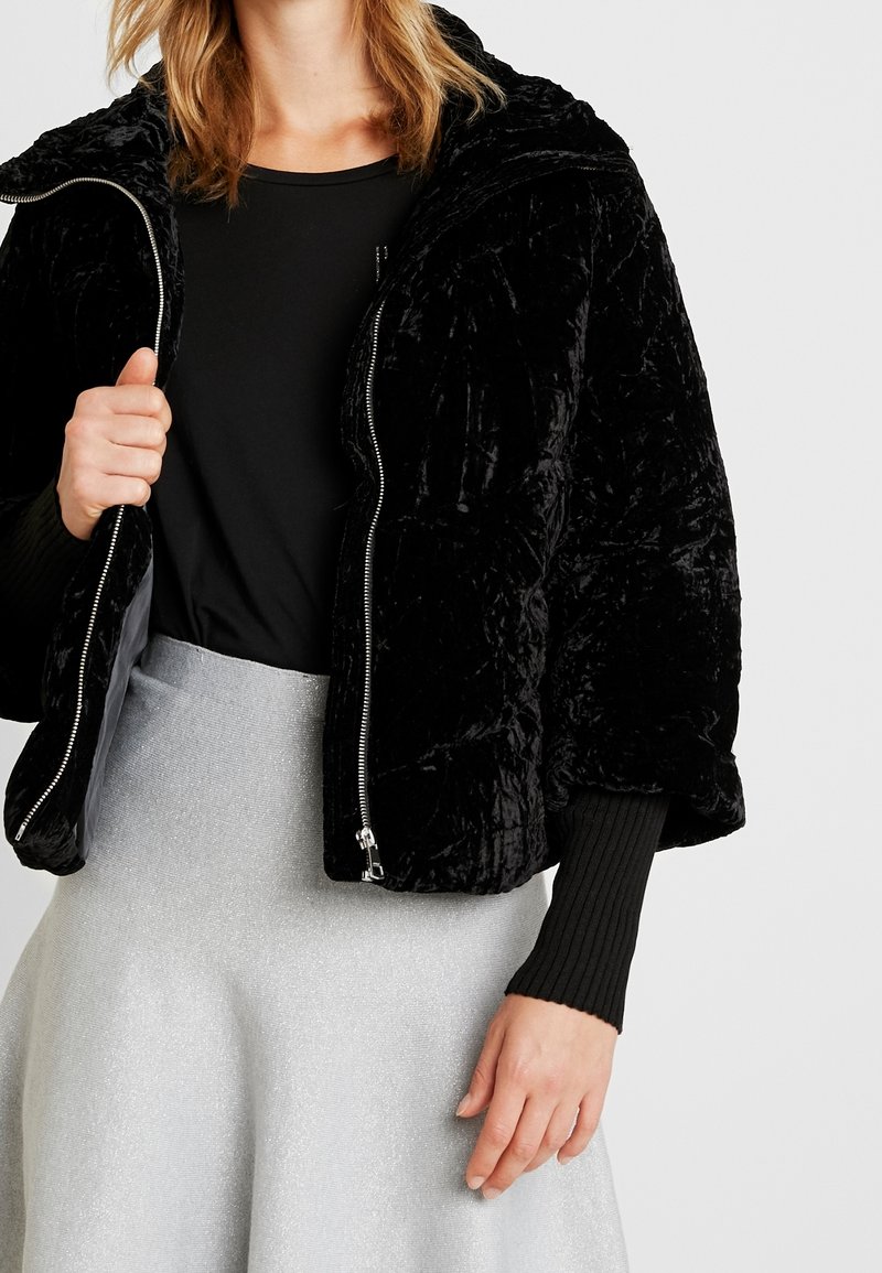 Femme portant une doudoune en velours noire par-dessus un top noir et une jupe texturée gris clair, debout devant un fond uni.