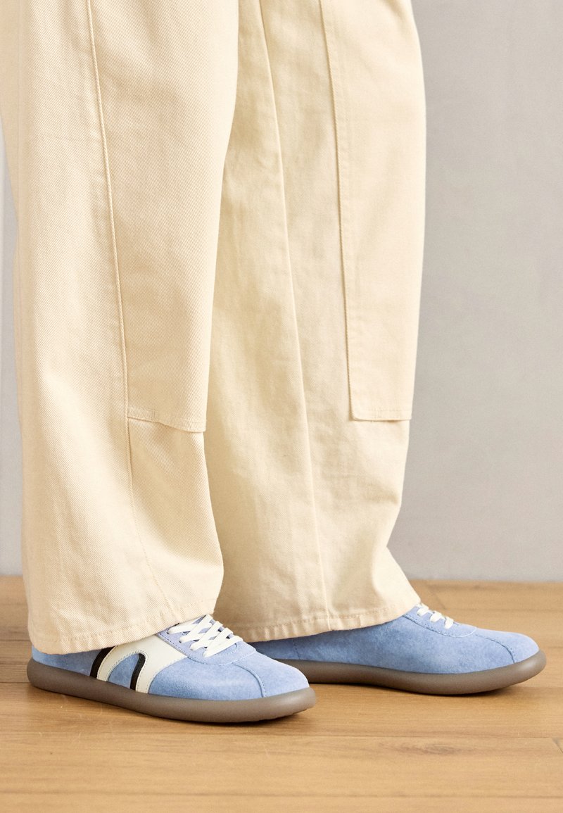 Zapatillas de ante azul claro con un diseño blanco, suela de goma gris y cordones blancos, colocadas al lado de pantalones de pierna ancha color crema.