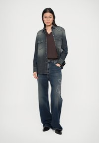 Γυναίκα που φοράει ξεθωριασμένο μαύρο τζιν πουκάμισο, σκούρο καφέ μπλουζάκι με V, φαρδιά κεντημένα τζιν και μαύρα παπούτσια, κρατώντας το ένα χέρι στην τσέπη.