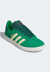Green suede sneaker with a textured white sole, featuring yellow three-stripe accents, green laces, and a padded green tongue with a red logo.
