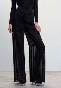 Femme portant un pantalon noir en pinstripe transparent à taille haute et jambes larges, avec un haut noir ajusté à manches longues et des talons ouverts argentés.