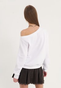 Sudadera blanca de hombros al aire con puños y dobladillo acanalados, combinada con una falda de mezclilla negra con bordes deshilachados y sutilezas en las costuras.