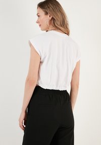Weißes, kurz geschnittenes Top mit kurzen Ärmeln und gerafftem Saum. Kombiniert mit schwarzen, elastischen Hosen mit seitlichen Taschen. Schlichte Stoffstruktur.