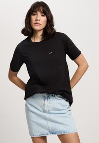 Czarna bawełniana koszulka z kieszenią i złotym akcentem, zestawiona z jasnoniebieską dżinsową mini spódnicą z zapięciem na guziki i delikatnym postrzępieniem.