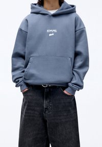Sudadera azul clara con un bolsillo delantero y un estampado gráfico blanco. Hecha de tela suave, con un corte relajado y puños acanalados. Combinada con jeans oscuros.