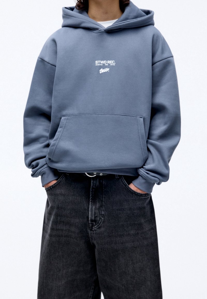 Sudadera azul clara con un bolsillo delantero y un estampado gráfico blanco. Hecha de tela suave, con un corte relajado y puños acanalados. Combinada con jeans oscuros.