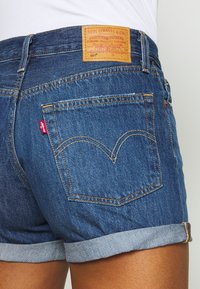 Shorts en denim avec un lavage bleu foncé, ourlet roulé, et une étiquette en cuir marron avec les détails de la marque. Logo rouge notable sur la poche arrière.