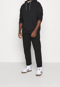 Black hoodie with a front pocket and gray drawstrings, paired with black trousers and white sneakers featuring black stripes and brown accents.