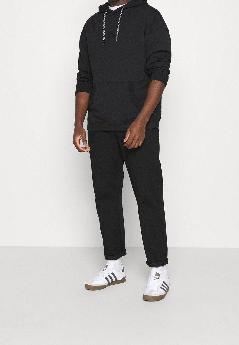 Black hoodie with a front pocket and gray drawstrings, paired with black trousers and white sneakers featuring black stripes and brown accents.