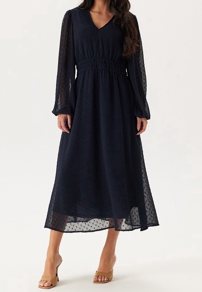 Femme portant une robe midi noire à manches longues en tissu transparent à pois et des talons ouverts couleur beige, debout sur un fond uni.