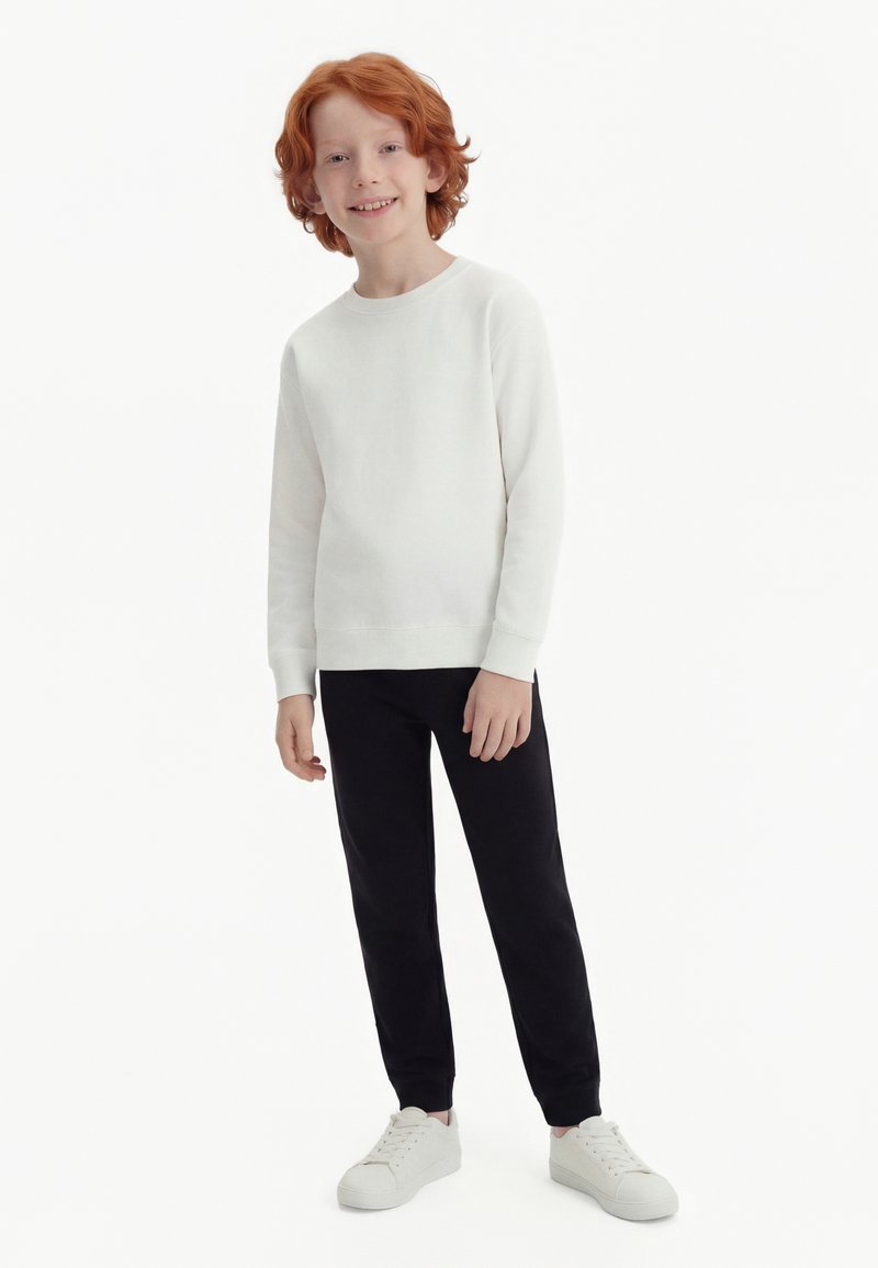 Rødenthåret dreng stående, iført en hvid langærmet skjorte, sorte bukser og hvide sneakers, smilende til kameraet.