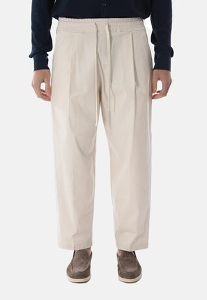 Uomo che indossa pantaloni beige a vestibilità rilassata con cintura regolabile a coulisse e pieghe, abbinati a una camicia blu scuro a maniche lunghe e mocassini in camoscio marrone.