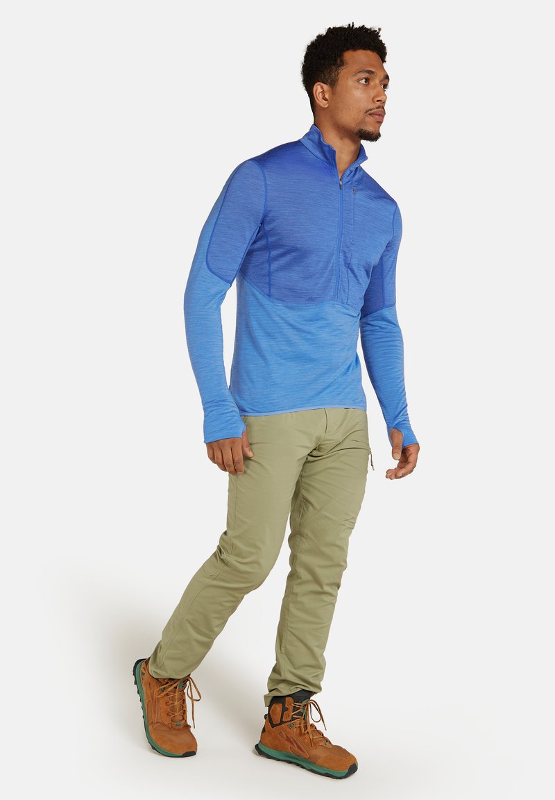 Top de manga larga azul con cremallera de un cuarto y diseño en degradado, combinado con pantalones cargo beige y botas de senderismo marrones. Cuenta con agujeros para los pulgares en las mangas.