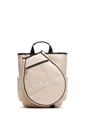 Kreemjas toonis kandekott musta ääre ja sangaga, esikülje lukuga taskuga, mis on kujult nagu tennisesaadetise kate, millel on kiri „lululemon“.
