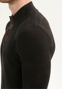 Detailní záběr muže oblečeného v tmavě hnědém žebrovaném pleteném svetru s opatřeným vysokým límcem a texturovanými detaily na ramenou.