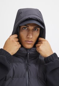 Črna pufasta jakna z visokim ovratnikom in elastičnimi manšetami, nosi se z ujemajočo kapo. Tkanina se zdi gladka in izolirana.