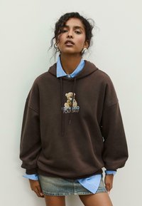 Brauner oversized Hoodie mit einem Bären-Graphic und dem Text „I ESPRESSO YOU’SELF“, übereinander getragen über einem hellblauen Hemd. Jeansrock. Weiche Textur.