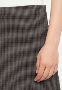 Podrobno prikazana roka, ki se naslanja na krilo v sivi in črni houndstooth vzorcu, oblečeno z belo majico.