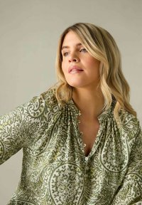 Blusa verde a fantasia con scollatura a V, caratterizzata da dettagli arricciati e piccoli bottoni, su un tessuto leggero. Texture morbida con un design floreale.