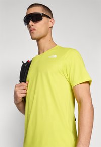 Camisa de manga corta verde lima con un patrón texturizado, que presenta un logo blanco. Gafas de sol negras y una mochila negra con un acento rojo son visibles.