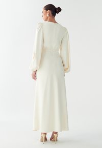 Vestido largo color crema con mangas abullonadas, cintura entallada y falda fluida y suave. La parte trasera tiene un cierre con cremallera sutil. Combinado con tacones nude.