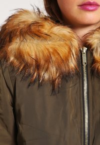 Olivgrön jacka med ett plysch-liknande fuskpäls-kant i nyanser av brunt och guld. Har en framfick med en dragkedja i guldton. Slät textur.