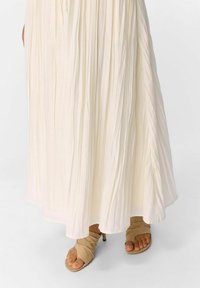 Falda maxi de crema ligera y plisada con un diseño fluido; combinada con sandalias de tacón beige que tienen una suave correa drapeada.