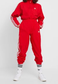 Röd träningsdräkt i lättviktsmaterial, med en jacka med dragkedja och vita ränder samt matchande byxor, dekorerad med Adidas-logotypen.