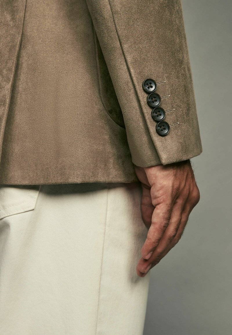 Blazer en daim marron avec trois boutons noirs sur les poignets, associé à un pantalon beige clair, présentant une surface texturée et des lignes épurées.
