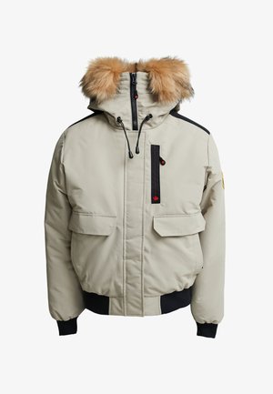 Beige winterjas met een afneembare bruine bontcapuchon, zwarte accenten, twee zijzakken, een ritszak op de borst en elastische manchetten.