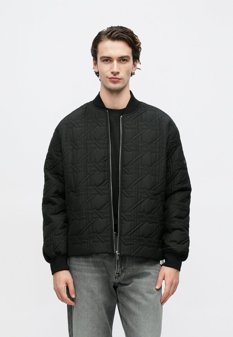 Νέος άνδρας που φοράει μαύρο καπιτονέ bomber μπουφάν με γεωμετρικό ράψιμο, μαύρο πουκάμισο από κάτω και γκρι τζιν, στέκεται μπροστά σε λευκό φόντο.