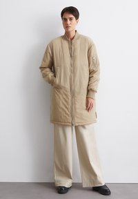 Beige lange jas met een rits aan de voorkant en ribgebreide manchetten, met zijzakken en een klein ritszakje op de mouw. Gecombineerd met beige wijde broek.