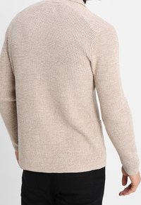 Beige stickad tröja med en texturerad design, långa ärmar och rund halsringning, med subtila sömnadsdetaljer på ryggen.
