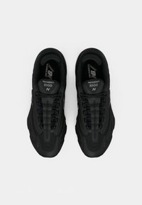 Baskets noires avec un empiècement supérieur texturé et respirant, un design à lacets et une marque New Balance bien en vue sur la languette et sur le côté.