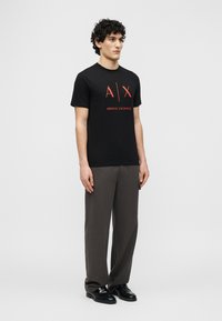 Νεαρός άνδρας όρθιος, φορώντας μαύρο t-shirt Armani Exchange με κόκκινο λογότυπο, σκουρόχρωμο γκρι παντελόνι με φαρδιά πόδια και μαύρα επίσημα παπούτσια, ουδέτερο φόντο.
