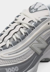 Baskets New Balance 1000 blanches et grises avec des panneaux en mesh, des lacets ondulés et le logo sur le côté et la languette, photographiées en gros plan.
