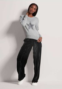 Grauer Pullover mit einem silbernen Sternenmuster und gerippten Bündchen, kombiniert mit schwarzen, samtigen weit geschnittenen Hosen, getragen mit weißen Sneakern.