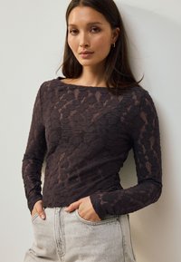 Tmavý krajkový top s dlouhými rukávy a výstřihem typu loďka, s texturovaným vzorem, nošený s světlými šedými džíny s vysokým pasem.