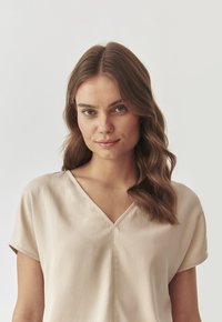 Beige satiini paita, jossa on V-mallinen pääntie ja lyhyet hihat, joka on kauniin sileä ja jossa on vain vähän ompelutietoja.