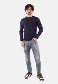 Maglione blu navy a maniche lunghe abbinato a jeans chiari strappati e sneaker marroni, con dettagli bianchi e un design classico.