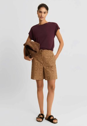 Donna con top viola senza maniche e pantaloncini beige con fantasia, che tiene una pochette marrone testurizzata, in piedi con sandali neri.
