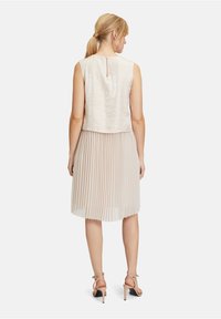 Faltenrockkleid mit einem hellbeigen Rock und einem strukturierten, kurzen Oberteil mit einem Schlüsselloch-Rückenausschnitt. Kombiniert mit nuden High-Heels.