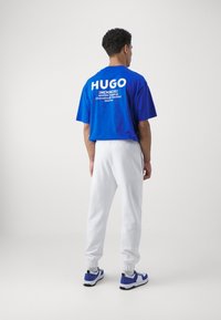 Blaues, übergroßes T-Shirt mit weißem "HUGO"-Logo auf dem Rücken, kombiniert mit weißen Jogginghosen und blauen sowie schwarzen Sneakern.