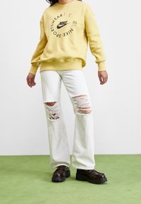 Žlutá mikina Nike s grafickým logem, doplněná o bílé roztrhané džíny s velkými trhlinami. Kombinováno s robustními černými botami na platformě.