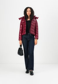 Bordeaux pufferjack met een glanzende afwerking, fitted ontwerp en capuchon. Gepaard met donkere flared jeans en een kleine zwarte handtas.
