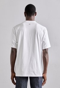 Camiseta de algodón blanca de corte relajado, con mangas cortas y un pequeño logo negro en la parte posterior del cuello. Textura suave, peso medio.