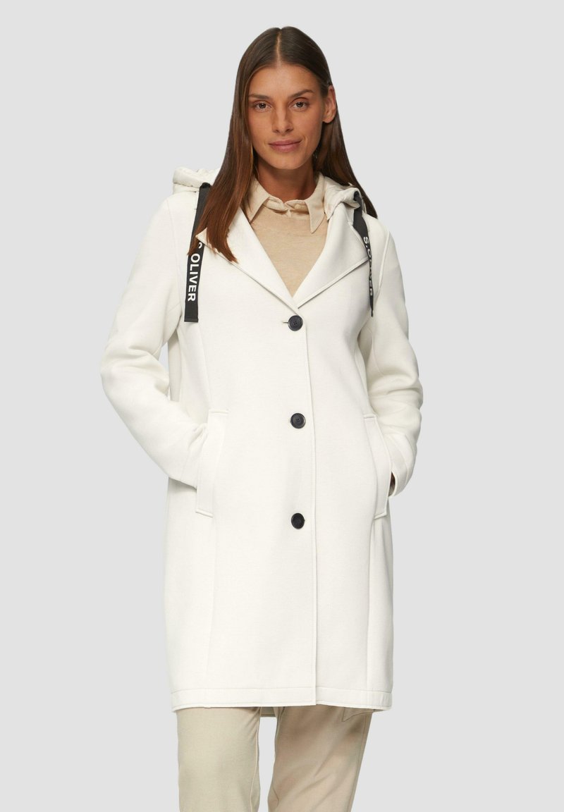 Cappotto lungo bianco con cappuccio, bottoni neri e tasche laterali. Presenta un colletto e cinghie nere in contrasto sul cappuccio. Texture liscia.